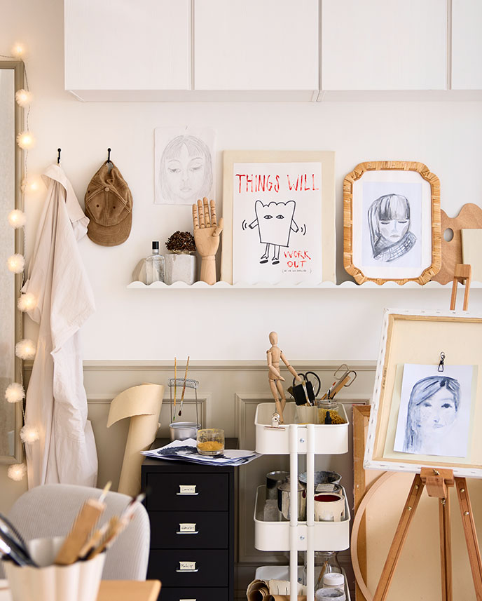 Una zona de manualidades en un dormitorio con una repisa para cuadros DAGGKAPRIFOL blanca en la que se exponen obras de arte y objetos decorativos en la pared.