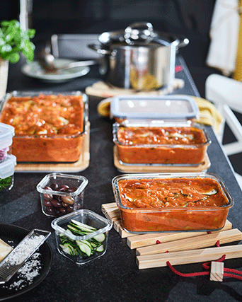 Recipientes de vidrio con tapas de bambú llenos de pasta con tomate sobre una encimera oscura junto a cuencos con aceitunas y calabacín.