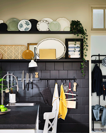 Cocina moderna con pared de azulejos negros, riel magnético para cuchillos, ganchos con paños amarillos y estantes con platos decorativos.