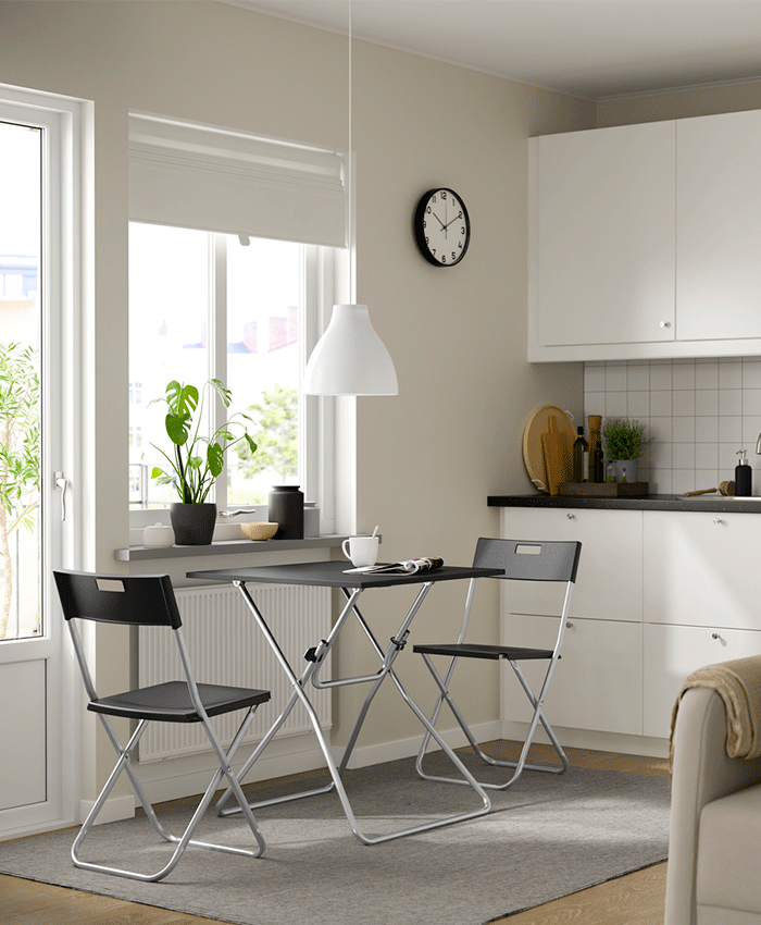 Small dining area in a bright kitchen featuring a black folding table and two matching chairs on a grey rug, positioned neatly under a window.