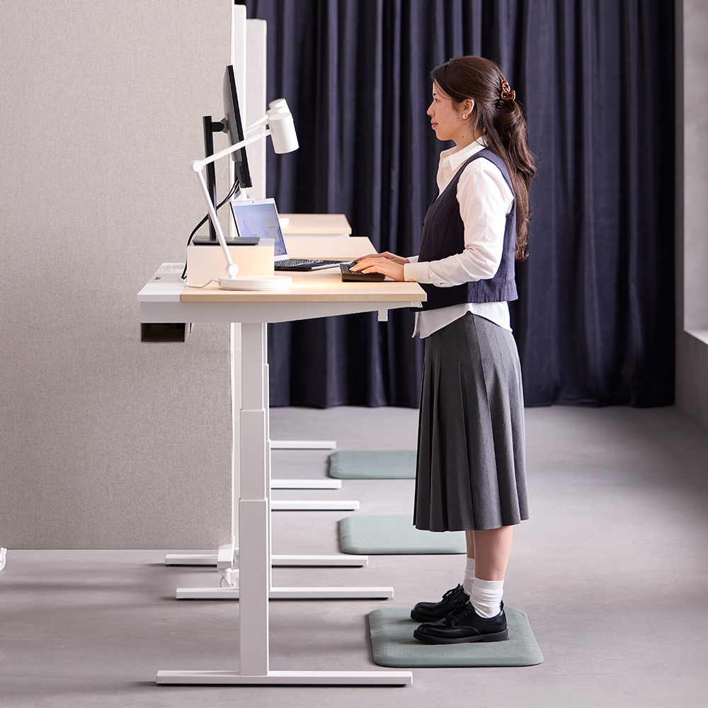 Office scene with a person standing while using a white MITTZON birch veneer sit/stand desk in high mode at a computer setup.