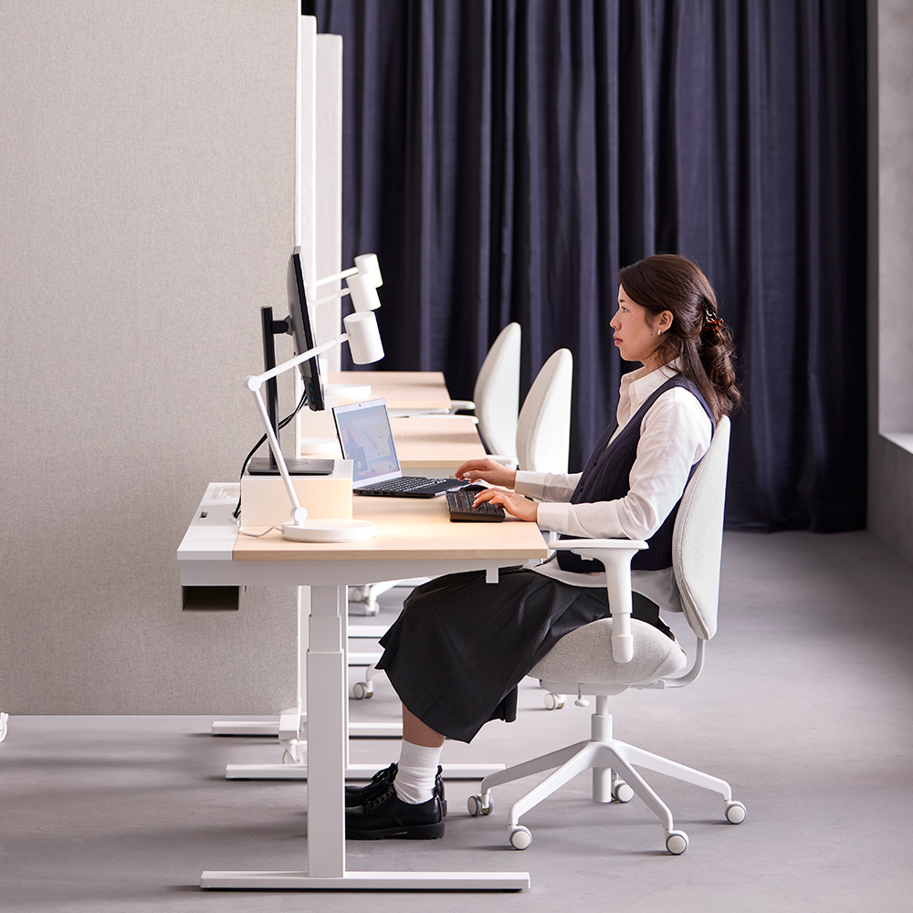 Office scene with a person using a white MITTZON birch veneer sit/stand desk on low mode, sitting by a computer setup.
