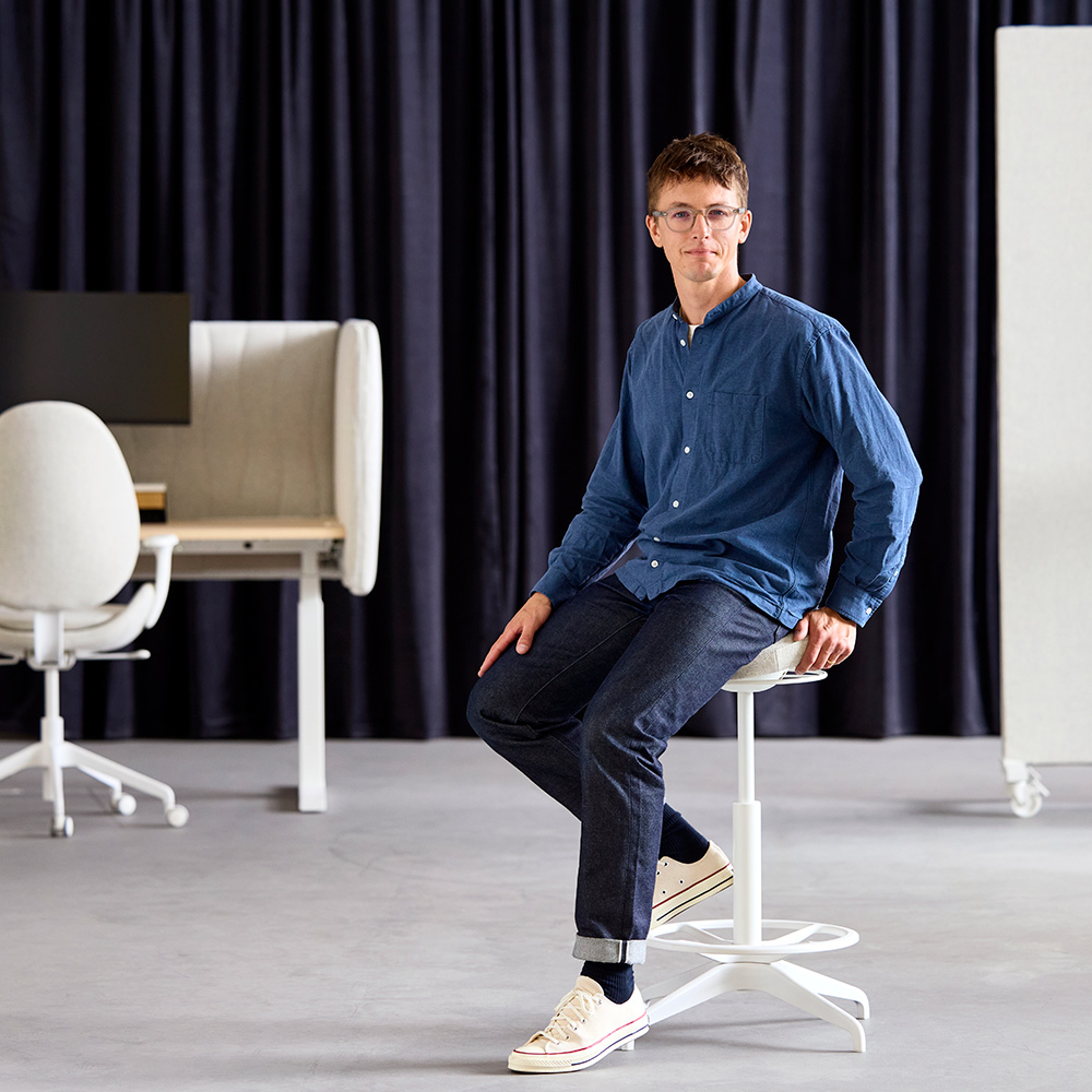 Philip Dilé, product developer, sits on a beige LIDKULLEN active sit/stand support, with a sit/stand desk in the background.