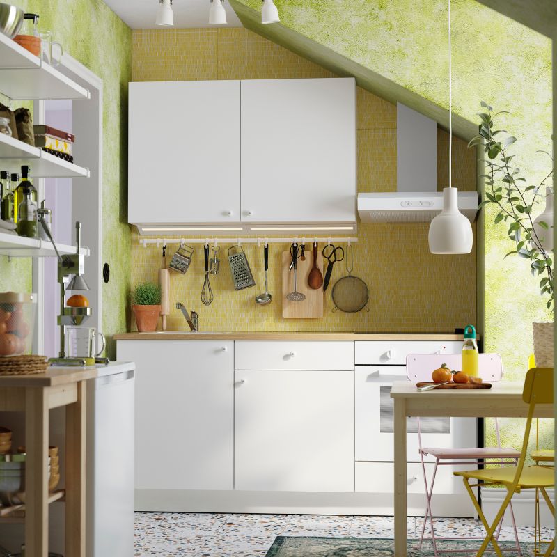 Cocina luminosa y compacta con gabinetes blancos, salpicadero amarillo, luz natural y utensilios y plantas colgantes.