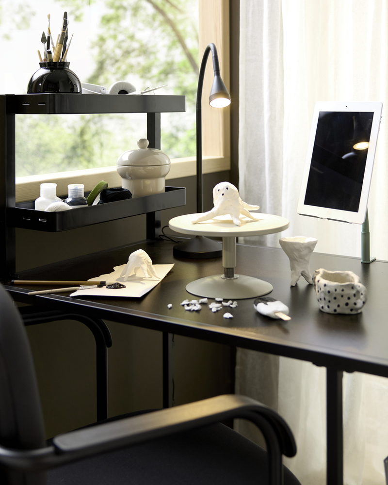 A close-up on a black-brown VITTSJÖ desk, with ongoing crafting, stationery and a lamp next to a digital tablet.