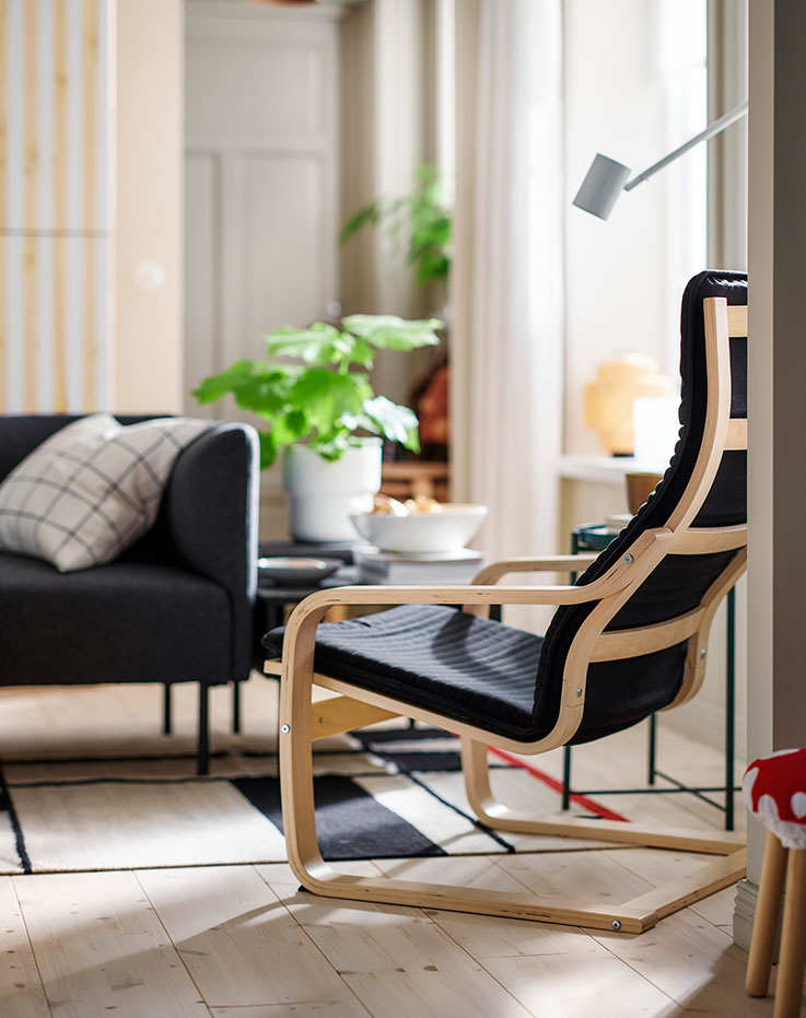 Una luminosa sala de estar con plantas frondosas y un sillón negro POÄNG fabricado con chapa de abedul, situado frente a un sofá LILLEHEM.