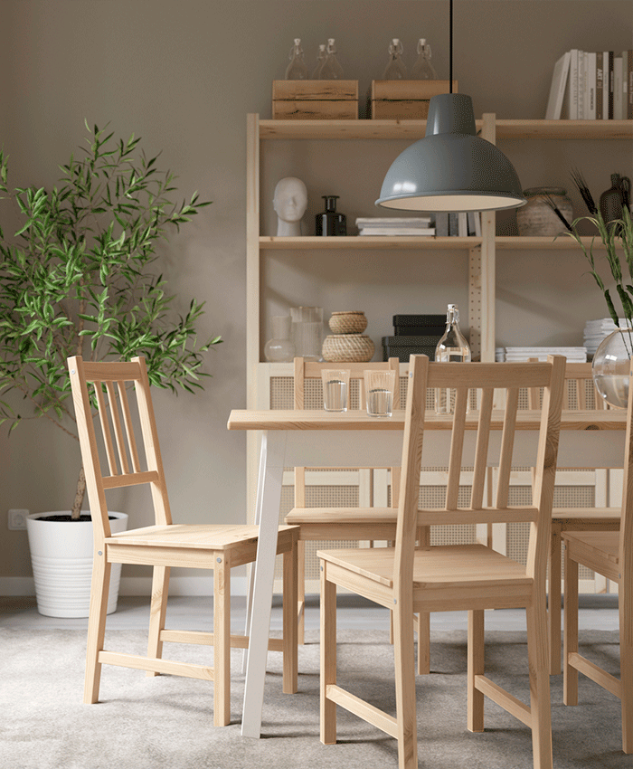 Light wood dining set with four chairs and a matching table in a bright room with a large plant, shelving unit, and a grey pendant lamp.