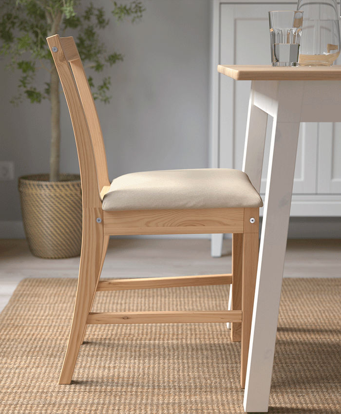 Side view of a light wood chair with a beige padded seat, placed next to a white-legged table on a natural fiber rug.