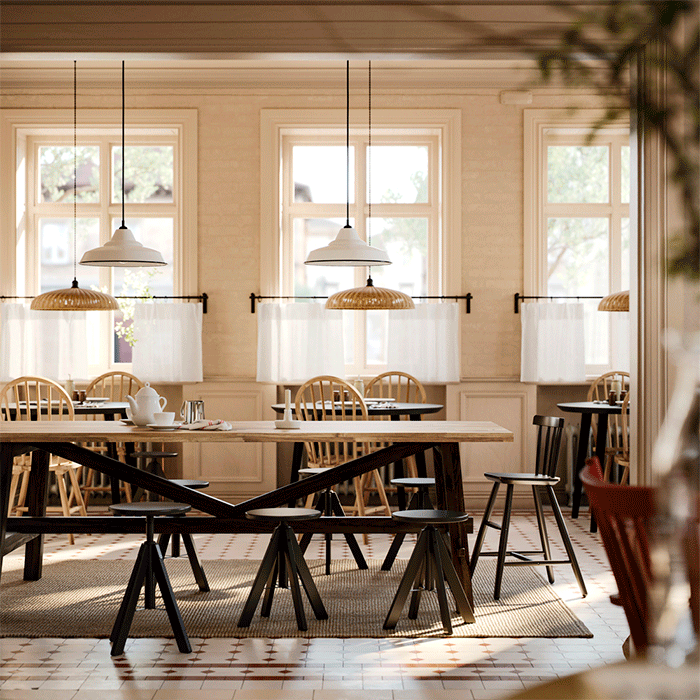 Spacious dining area with several wooden tables, black stools, and spindle chairs under warm pendant lighting near large windows with white curtains.