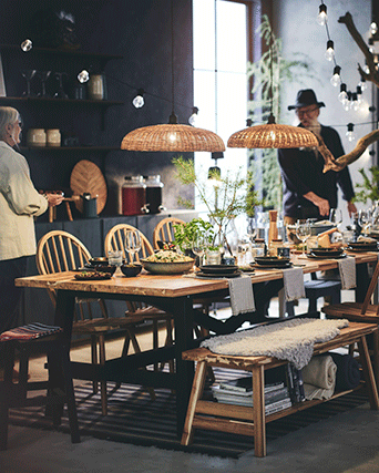 Long wooden dining table set for a meal with black legs, matching benches with faux fur, and two large woven pendant lamps hanging above.