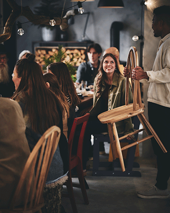 A man brings an extra light wood chair to a crowded, festive dinner table lit by string lights in a rustic, warm interior.