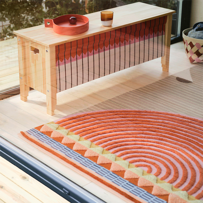 Bright room featuring a wooden bench with a patterned textile front, a round textured rug with orange circles, and a glass door to a patio.