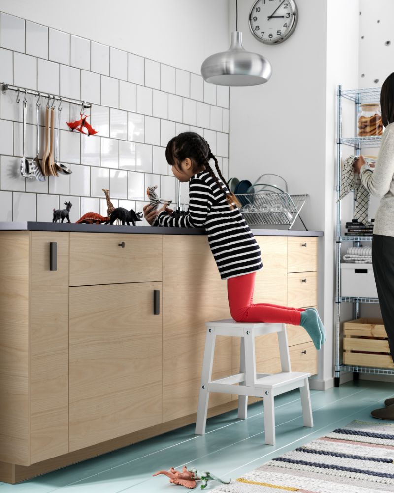 Niña juega con figuras en una cocina de gabinetes de madera clara y azulejos blancos, subida a un taburete blanco.