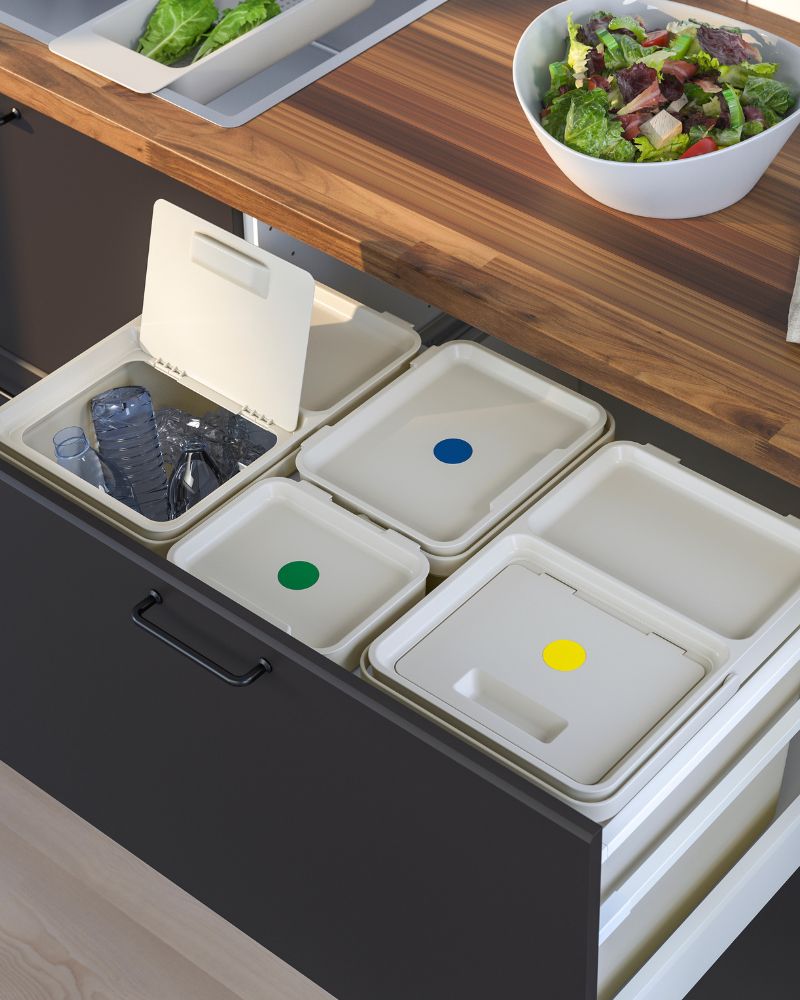 Cajón de cocina oscuro con botes de basura de reciclaje blancos y coloridos, bajo una encimera de madera.