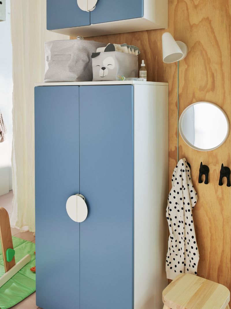 Un armario SMÅSTAD azul contra una pared de madera con dos cajas de almacenamiento con cosas de bebé encima y una lámpara de pared al lado. 