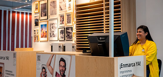 Smiling employee in a framing shop with a photo gallery wall and a light wood counter.