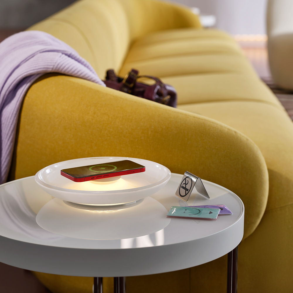Junto a un sofá verde amarillo hay una mesa blanca que sostiene un cargador inalámbrico VÄSTMÄRKE con forma de dona, iluminado por tarjetas de acceso del hotel.