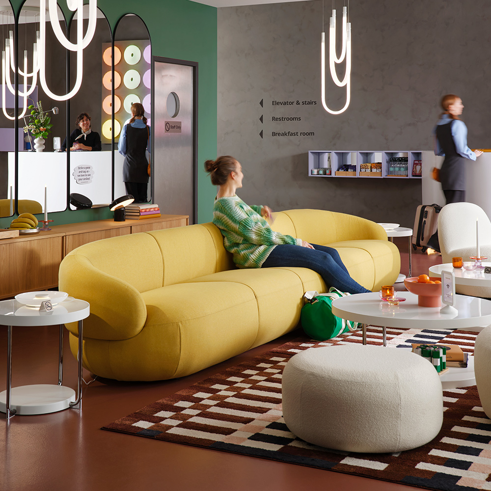 Una alfombra MARKBLÅMME de pelo corto con un patrón multicolor/cuadriculado se encuentra en un salón de hotel moderno que tiene huéspedes en el sofá y en la recepción.