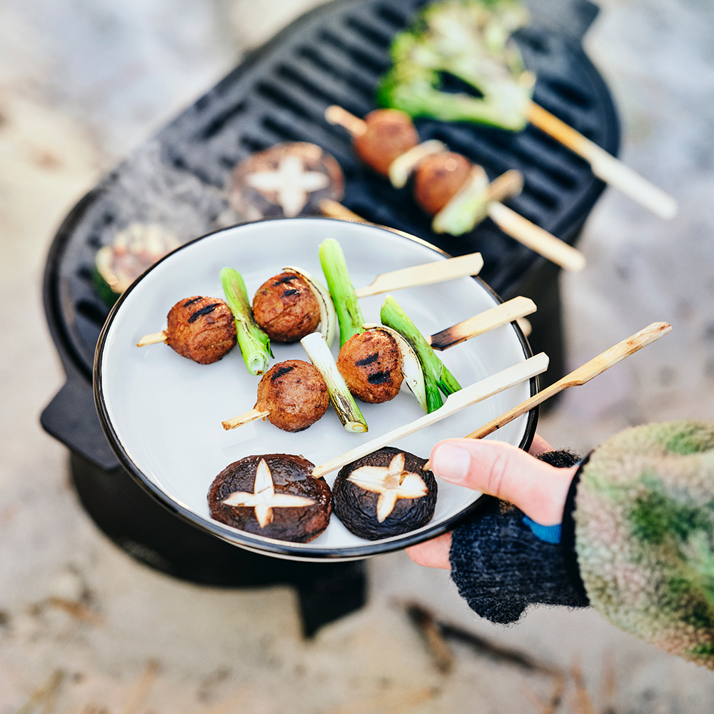 Una mano sostiene un plato SOLUPPGÅNG con brochetas de champiñones y albóndigas vegetales, cerca de una barbacoa SOLUPPGÅNG.