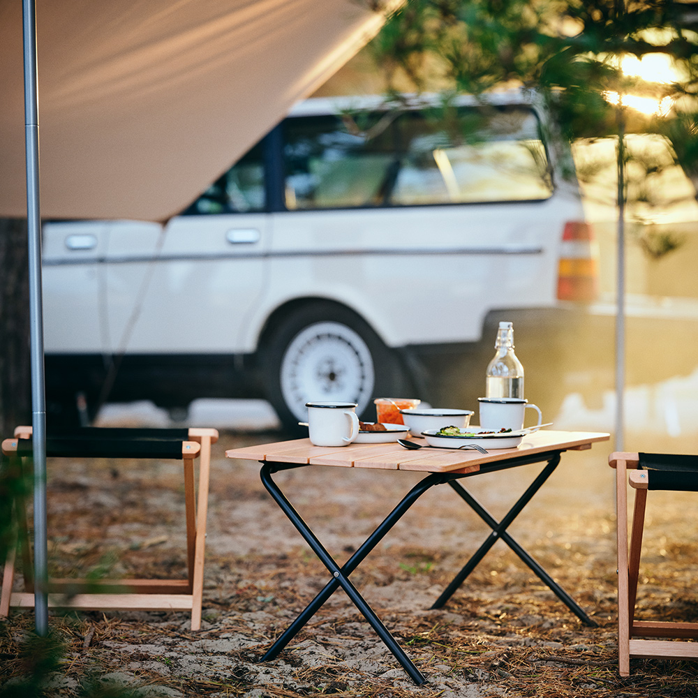 Una mesa plegable SOLUPPGÅNG con platos y tazas SOLUPPGÅNG encima está colocada bajo una lona, cerca de un coche.