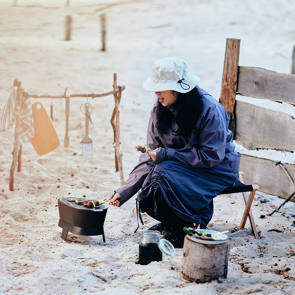Una persona que lleva un sombrero SOLUPPGÅNG está asando brochetas en una barbacoa SOLUPPGÅNG, en una playa de arena.