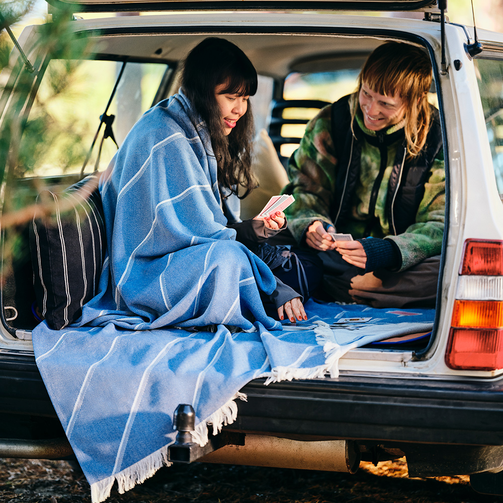 Una pareja sonríe y juega a las cartas en la parte trasera de un coche; una de ellas tiene una manta SOLUPPGÅNG envuelta alrededor de los hombros.