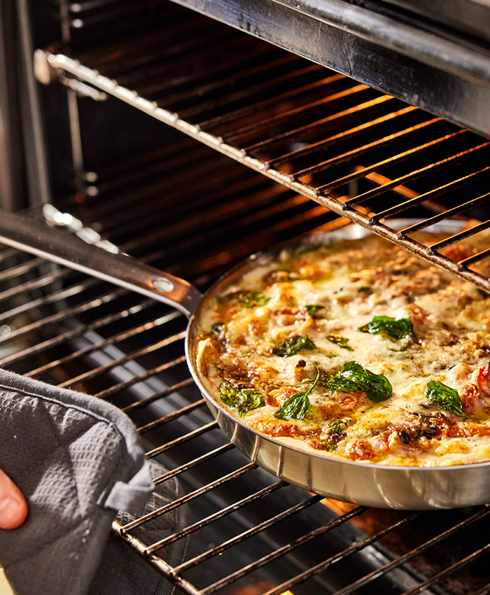 Frying pan with gratin food and spinach leaving the oven, held by a hand with an oven mitt.