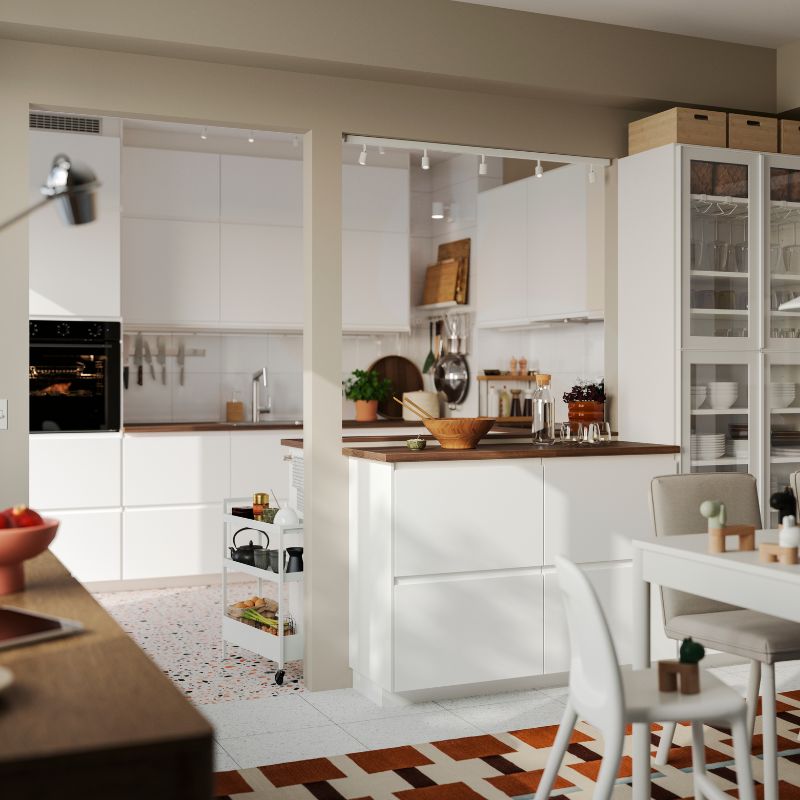 La luminosa cocina cuenta con frentes VOXTORP en blanco mate y una isla con una mesa de comedor blanca en primer plano.