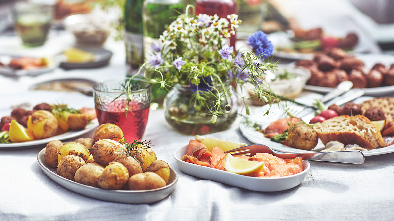 Mesa al aire libre con patatas asadas, salmón, albóndigas y un jarrón con flores silvestres, con el mar de fondo.