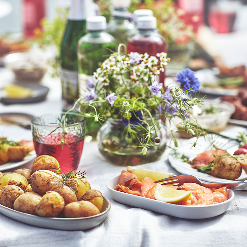 Mesa al aire libre con patatas asadas, salmón, albóndigas y un jarrón con flores silvestres, con el mar de fondo.