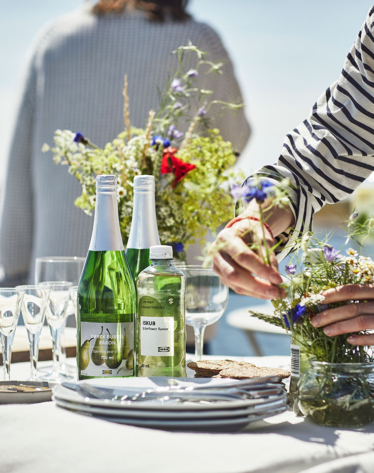 Mesa con botellas de bebida espumosa de pera y agua de saúco junto a platos de comida durante una reunión al aire libre junto al agua.