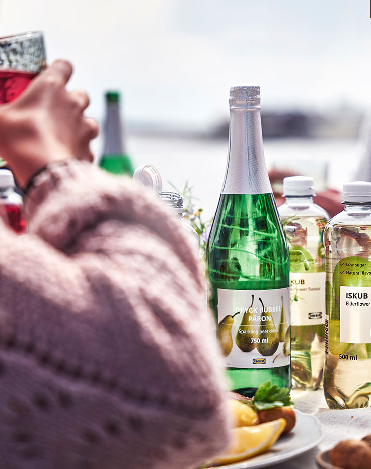 Mesa con botellas de bebida espumosa de pera y agua de saúco junto a platos de comida durante una reunión al aire libre junto al agua.