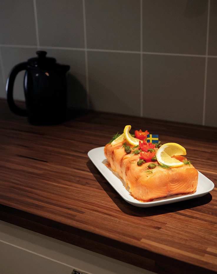  Plato de salmón decorado con rodajas de limón y eneldo sobre una fuente blanca en una encimera de madera oscura junto a una tetera negra.