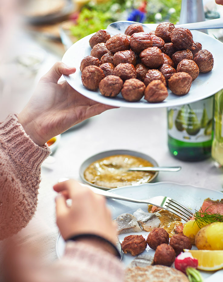 Alguien con jersey de punto rosa sostiene un plato de albóndigas sobre una mesa llena de platos variados y patatas.