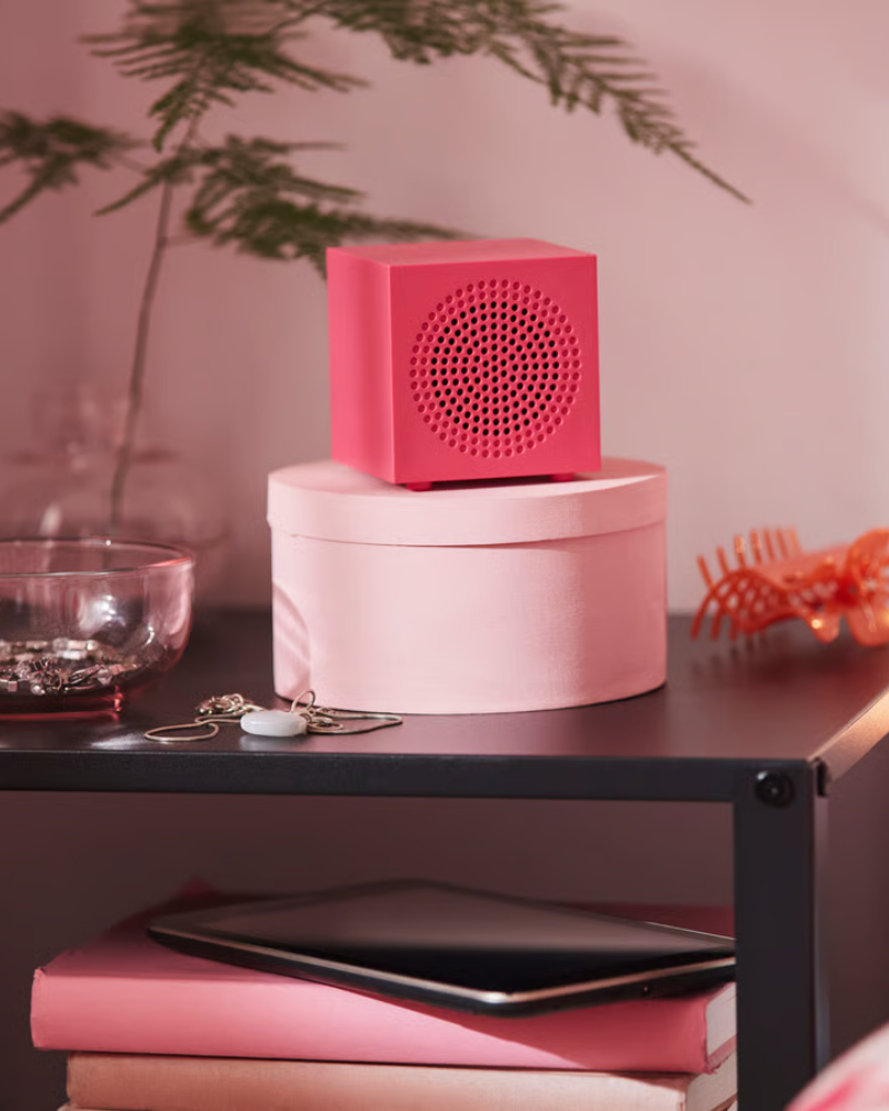 A close-up of a pink KALLSUP portable Bluetooth speaker, placed on a black bedside table together with decorative items.