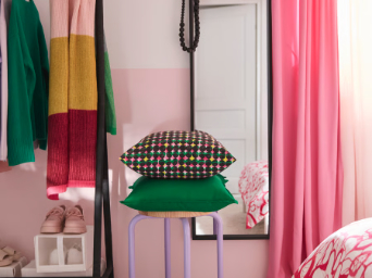 A bedroom with a pink/green ÄLVSJÖSKOGEN cushion cover placed on a green GURLI cushion cover stacked on a stool.