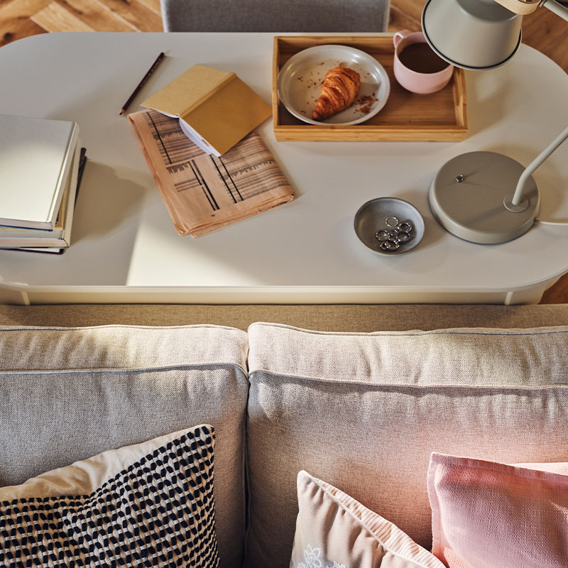 A tray with food and coffee, a lamp, a book and a newspaper stand on a ROSENTORP desk behind an EKHOLMA 2-seat sofa bed.