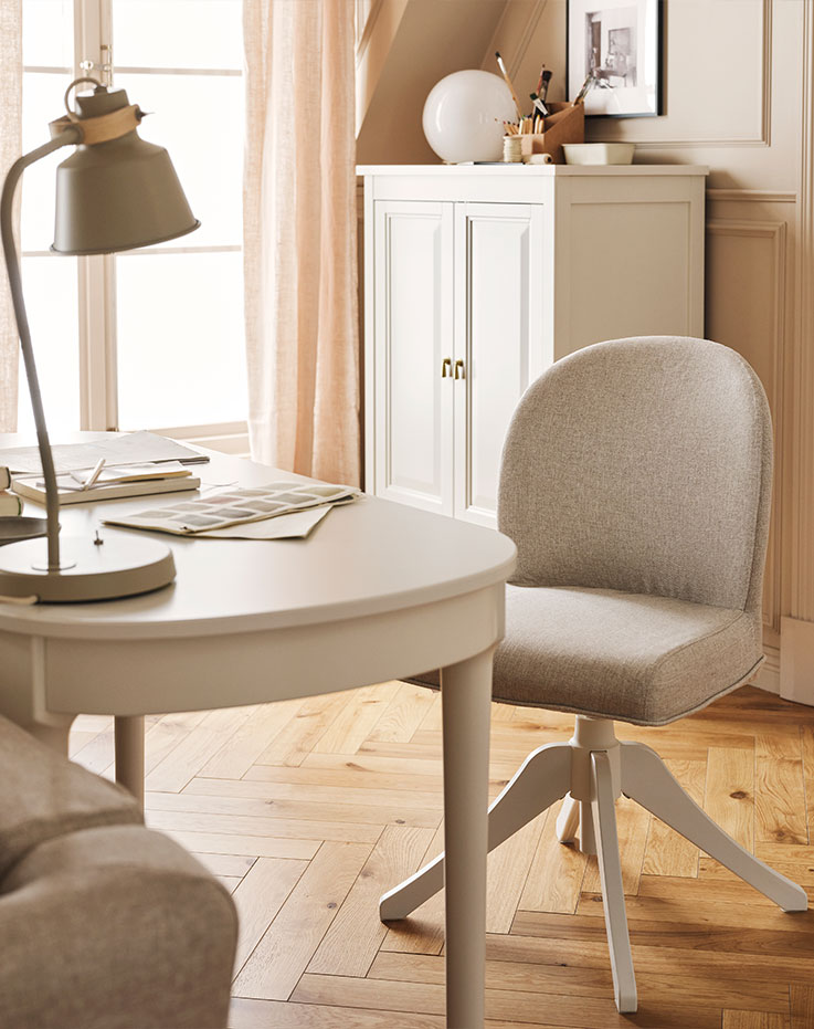A white ROSENTORP desk and a ROSENTORP swivel chair stand behind a sofa near an off-white FÄRJKARL cabinet in a living room.
