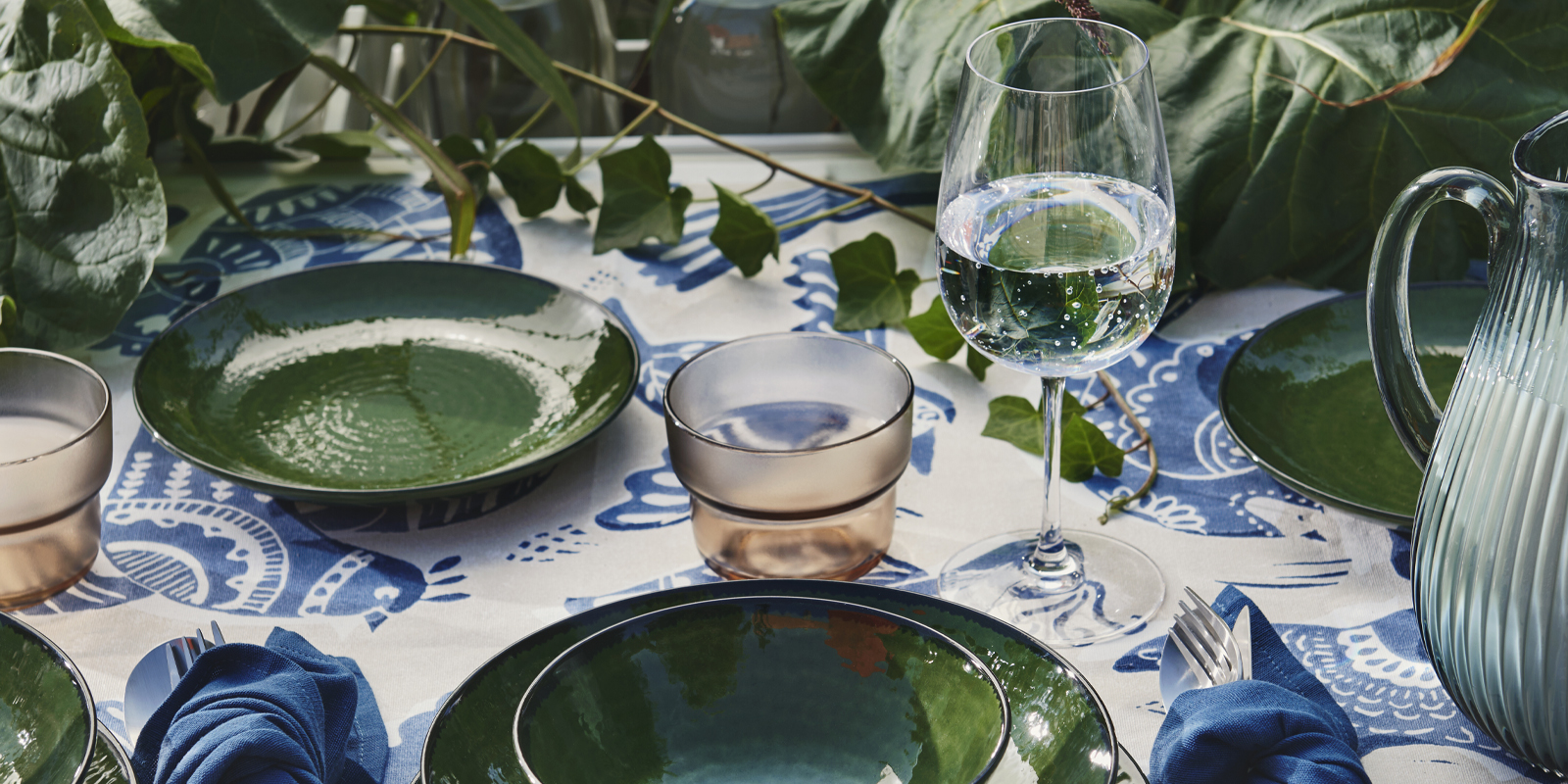 Una mesa de comedor al aire libre con un cuenco verde PELARKAKTUS junto a cristalería y plantas sobre un mantel floral.