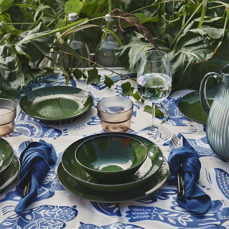 Una mesa de comedor al aire libre con un cuenco verde PELARKAKTUS junto a cristalería y plantas sobre un mantel floral.