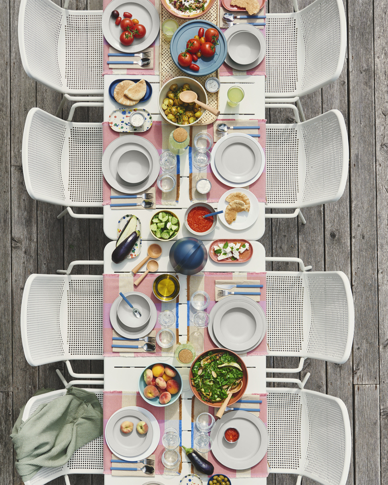 Una mesa de comedor al aire libre para ocho personas está puesta con platos SIMKRABBA de color verde grisáceo, y alrededor hay platos y vasos en tonos pastel.