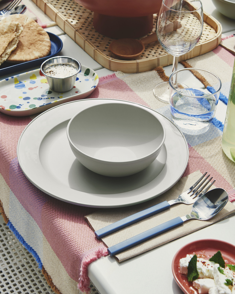 Una mesa de comedor al aire libre con un plato SIMKRABBA de color verde grisáceo, con diversos platos y vasos dispuestos a su alrededor.