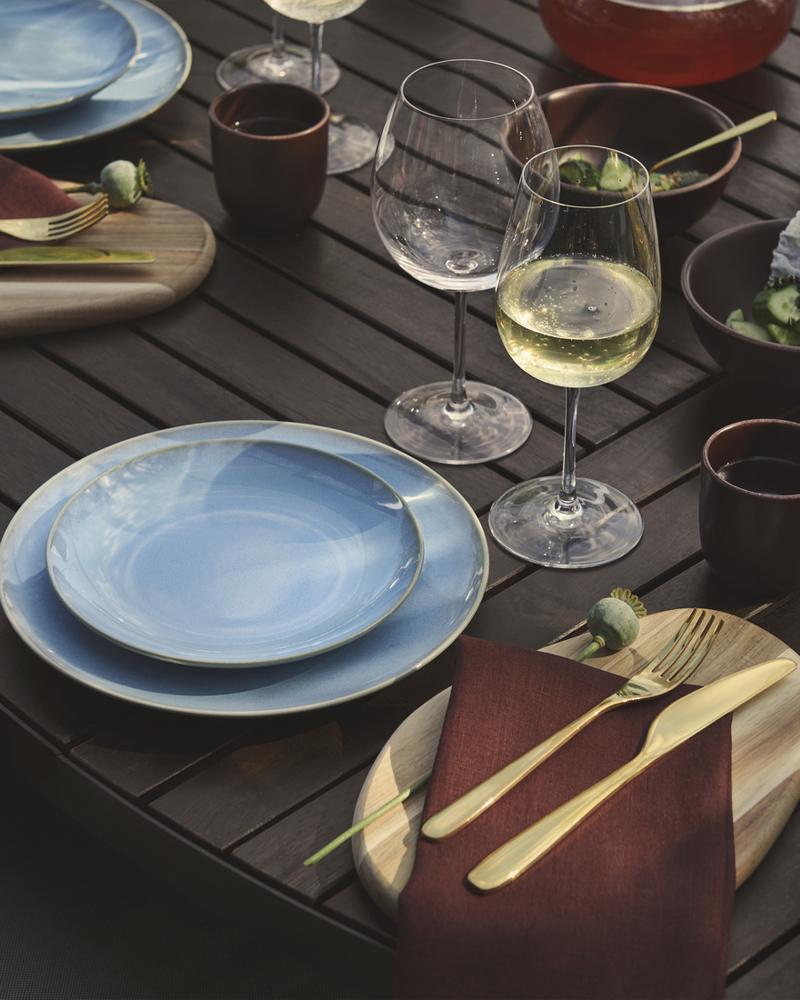 Una mesa de comedor al aire libre con vajilla FÄRGKLAR azul claro, vasos transparentes, cubertería dorada y bandejas de madera.