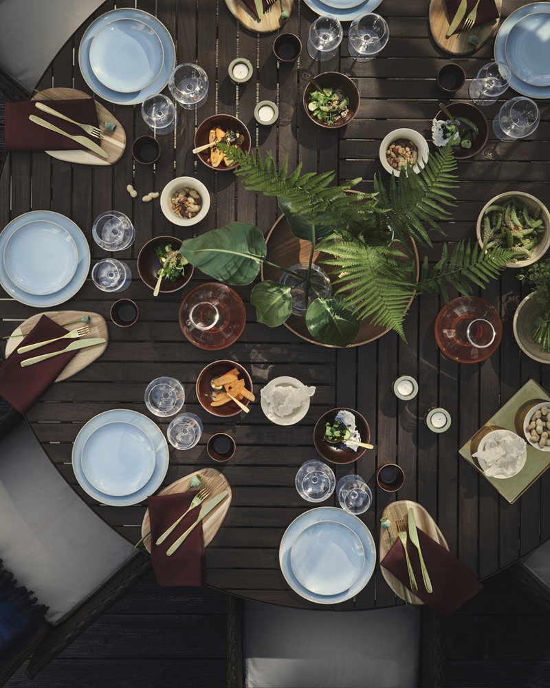 Una mesa de comedor al aire libre con una mesa redonda VITTSKÄR gris oscuro, decorada con vajilla FÄRGKLAR azul claro y plantas.