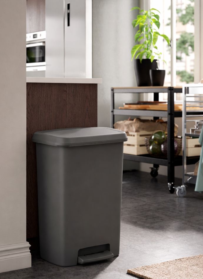 Bote de basura gris con tapa metálica en una cocina moderna con carrito auxiliar y planta verde.