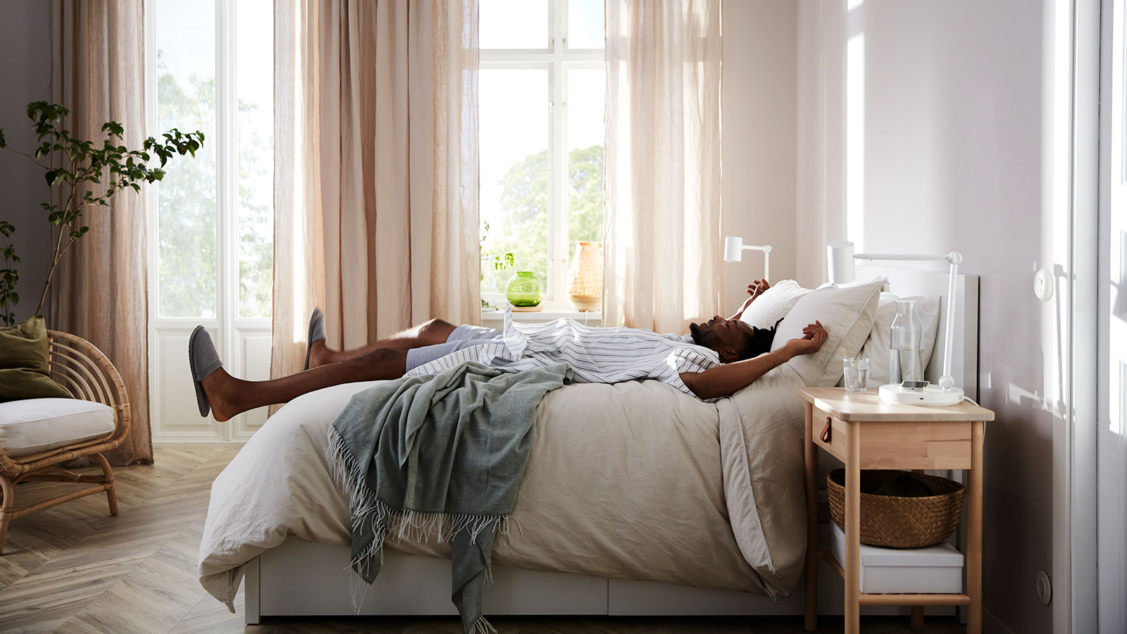 Imagen de un dormitorio luminoso con cortinas beige en vista lateral, con un hombre tumbado como si acabara de saltar y se hunde entre sábanas y colchas mullidas, la cama tiene aspecto de pino y estilo moderno.