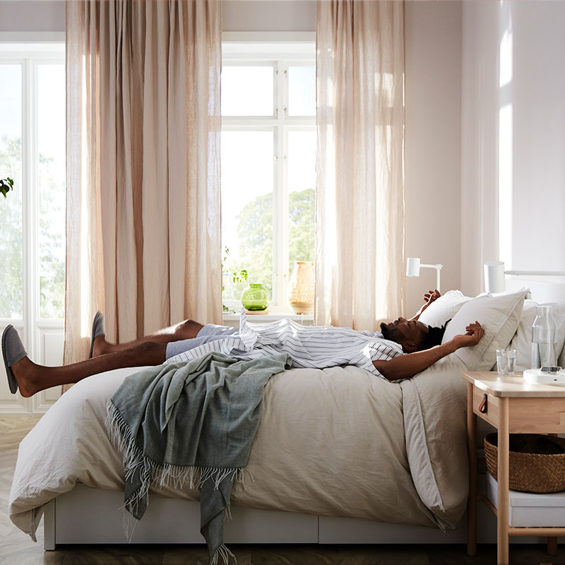 Imagen de un dormitorio luminoso con cortinas beige en vista lateral, con un hombre tumbado como si acabara de saltar y se hunde entre sábanas y colchas mullidas, la cama tiene aspecto de pino y estilo moderno.