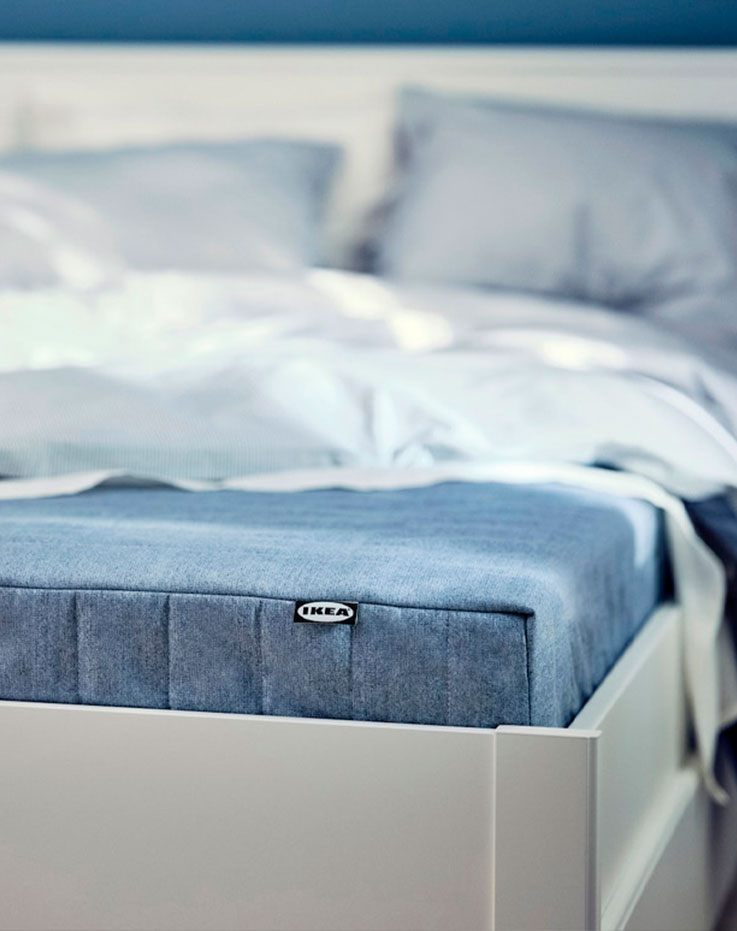 Esquina de una cama blanca de estilo moderno, con el colchón visible en azul y una colcha blanca encima.