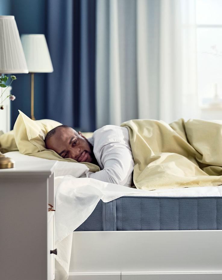 Una persona durmiendo cómodamente en una cama de estilo moderno, se ve el colchón azul, al fondo la cortina azul y blanca está desenfocada.