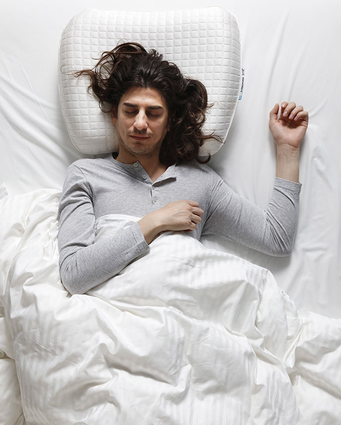 Persona tumbada boca arriba, sobre edredones blancos y con una almohada ortopédica.